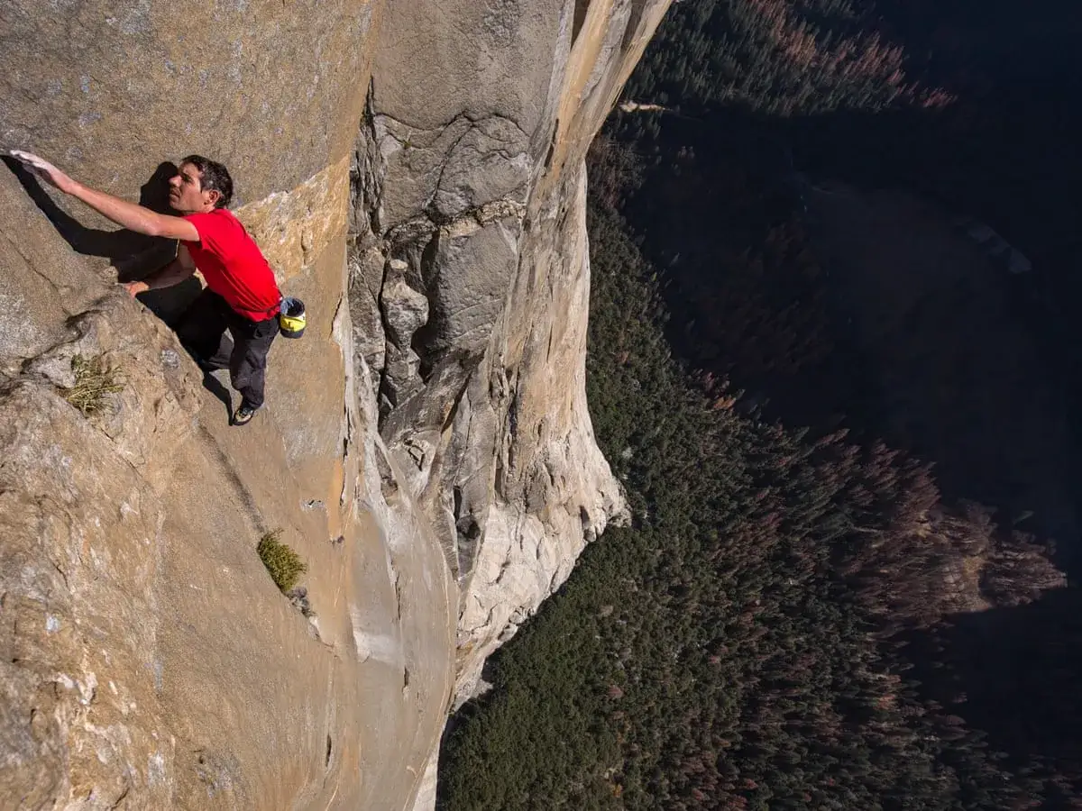 Alex Honnold klettert free solo den El Capitan. Ein Mann in Rot, der die Felswand bezwingt, mit einem tiefen Abgrund voller Bäume unter ihm.