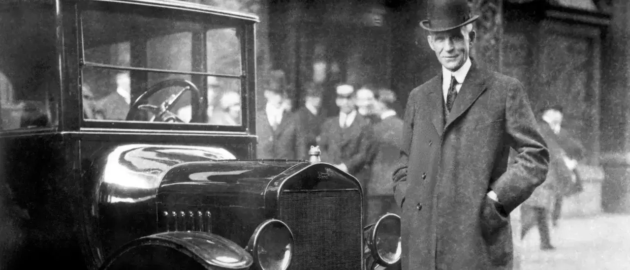 Henry Ford, właściciel Forda, stoi obok swojego samochodu w meloniku i płaszczu.