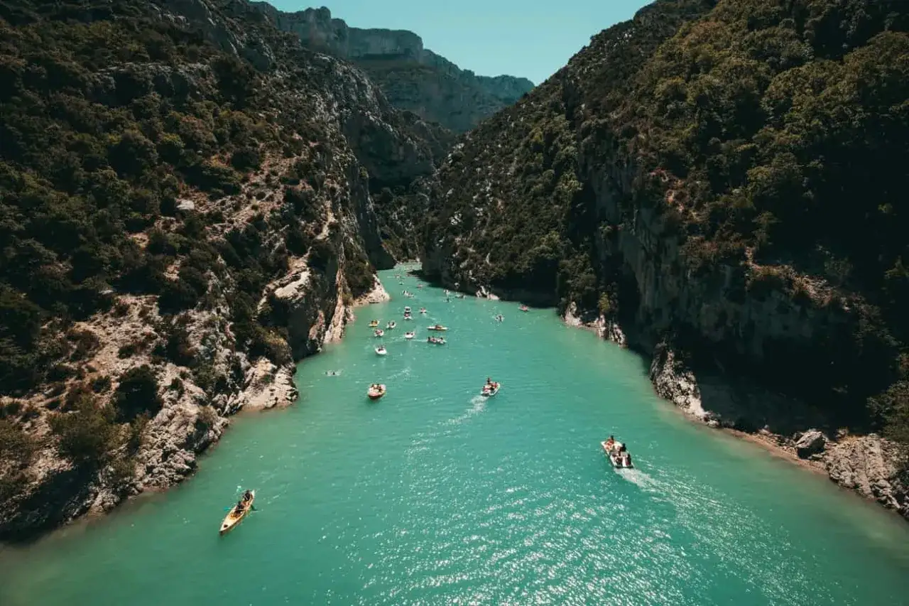 Un río turquesa serpentea entre acantilados verdes en Francia. Familias y niños disfrutan de un día soleado en kayaks y barcas.