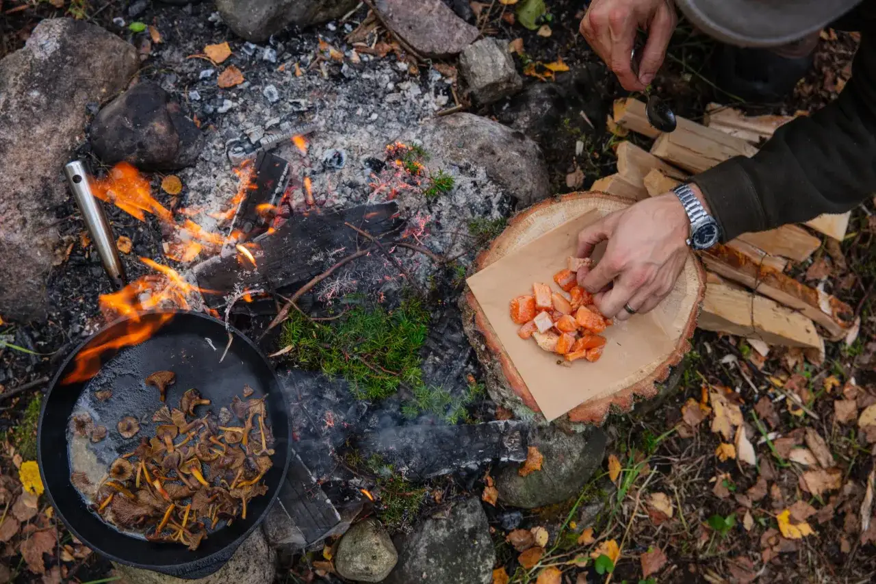 En person förbereder mat över öppen eld. Kantareller steks i en gjutjärnspanna medan tärnat kött läggs på en träbit. En smak av det ätbara landet.