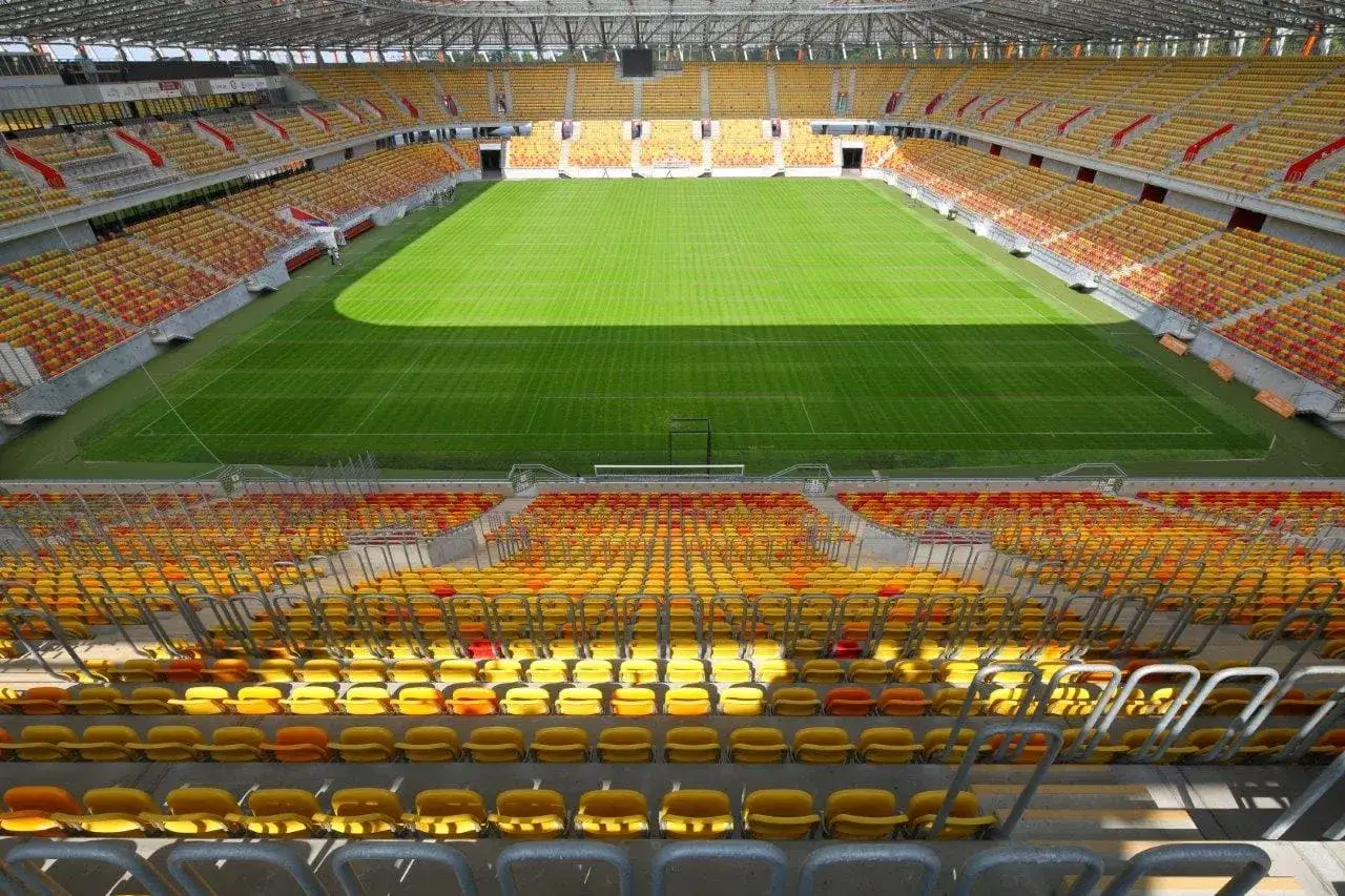 Pusta trybuna stadionu Jagiellonii, z żółtymi i czerwonymi siedzeniami, czeka na kibiców. Pojemność stadionu robi wrażenie.
