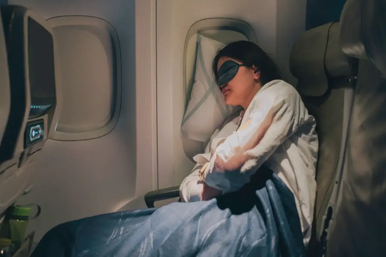 Frau schl&auml;ft im Flugzeug mit Schlafmaske, Decke und Kissen. Sie k&auml;mpft gegen den Jetlag von Asien nach Deutschland.