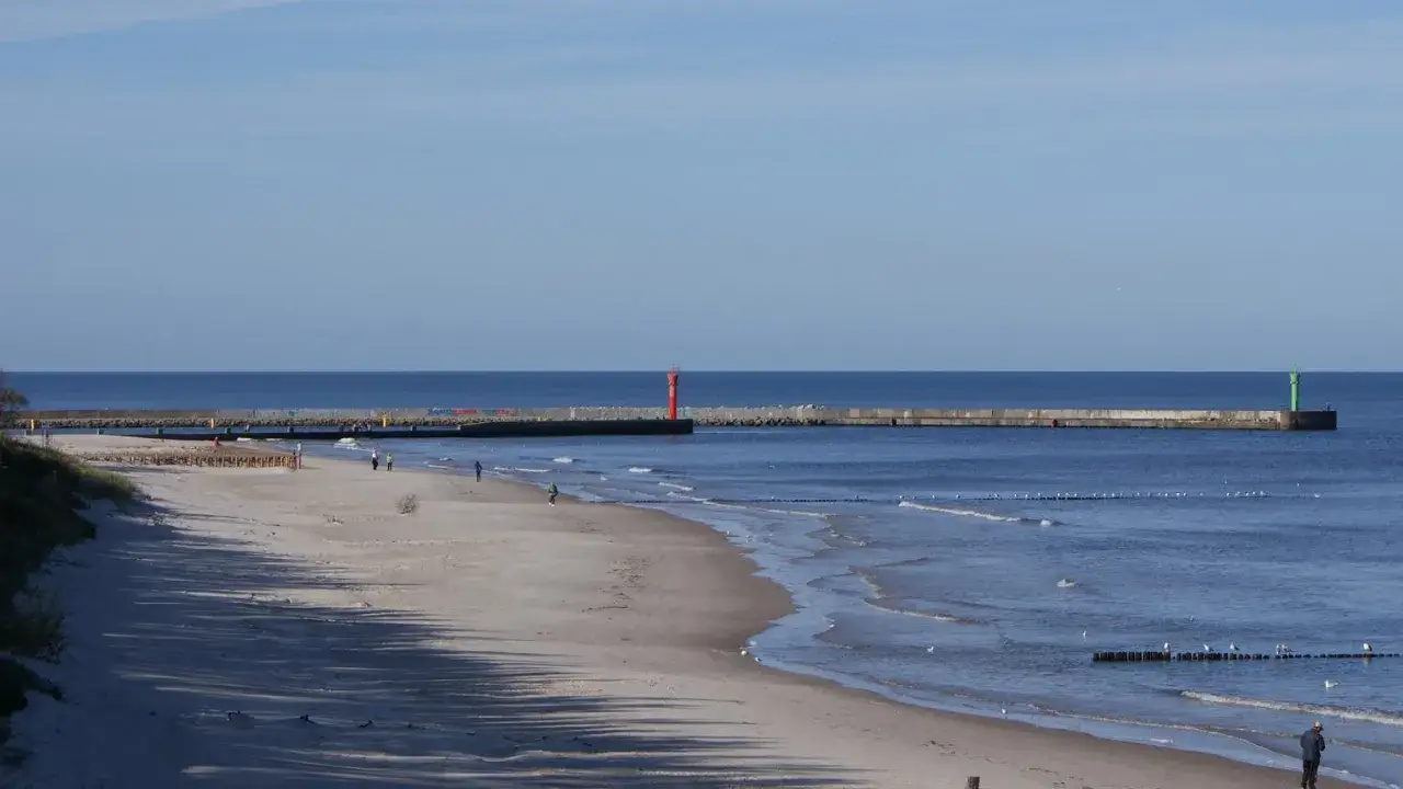 Plaża w Mrzeżynie z falochronem i latarniami morskimi. Spacerowicze cieszą się spokojem, a fale łagodnie wpadają na brzeg. Mrzeżyno atrakcje to idealne miejsce na relaks.