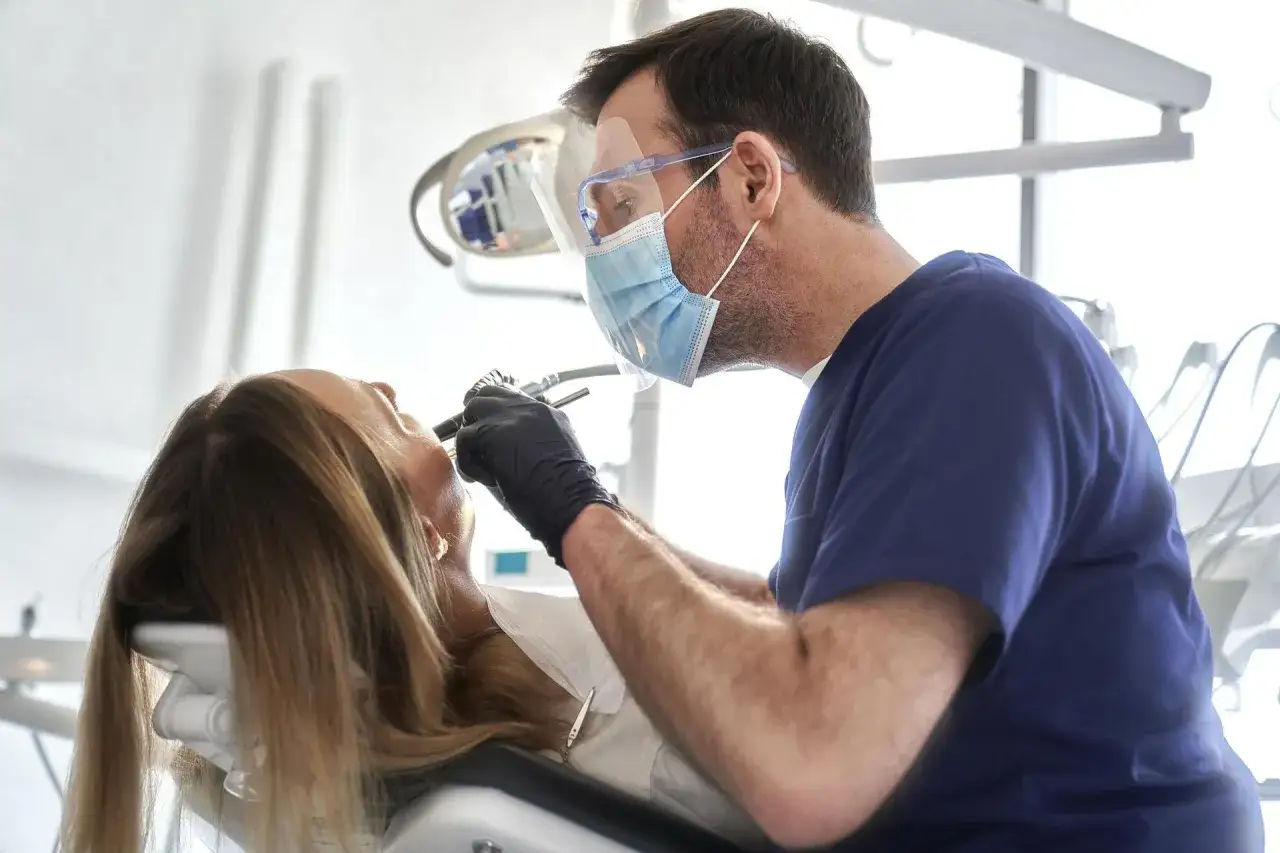 Dentysta w masce i przyłbicy wykonuje zabieg. Zastanawiasz się, ile kosztuje wyrwanie zęba?