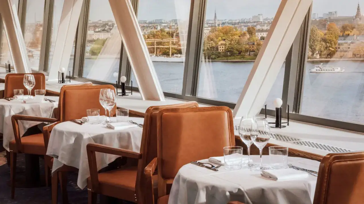 Eleganta bord med vita dukar och l&auml;derkl&auml;dda stolar i restaurang med utsikt &ouml;ver vatten och stad. Perfekt f&ouml;r gondolen recensioner.