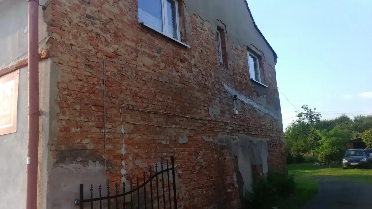 Pękające ściany starego domu z cegły, z widocznymi oknami i rynnami. W tle zieleń i samochody.