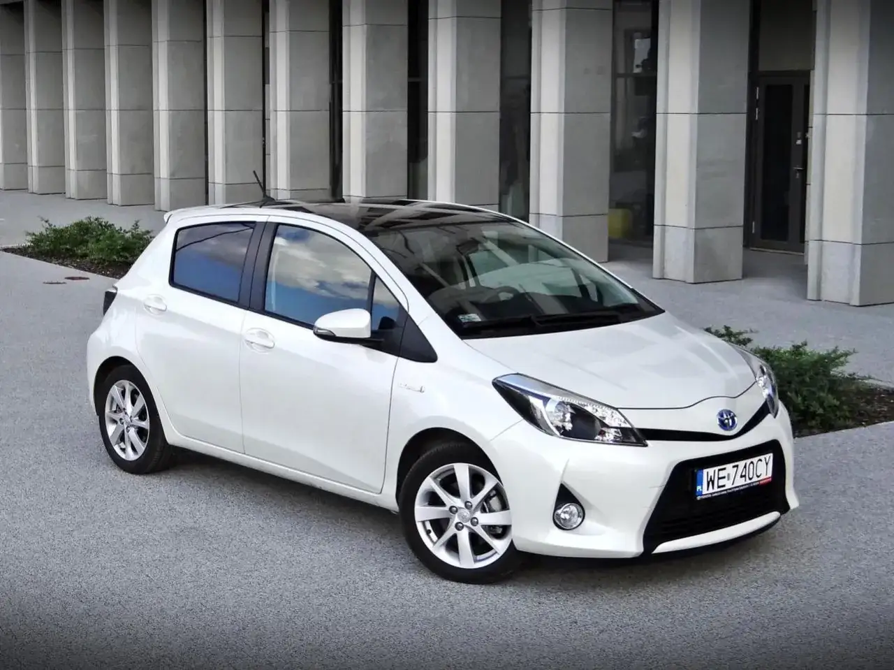 Biała Toyota Yaris Hybrid z czarnym dachem zaparkowana przed nowoczesnym budynkiem.