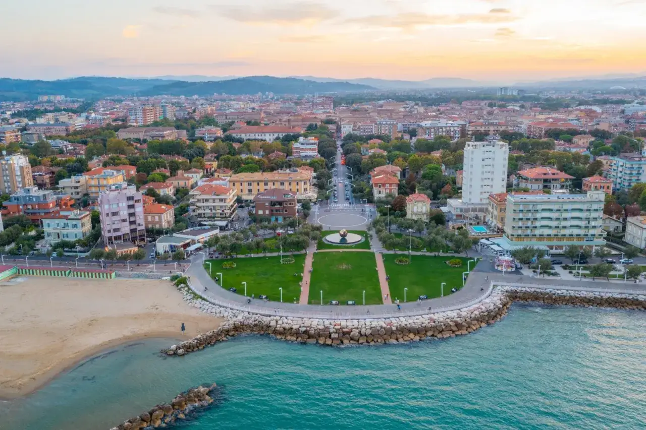 Vista aerea del **Pesaro lungomare** con la spiaggia, il mare turchese, un parco verde curato e la città che si estende all'orizzonte al tramonto.