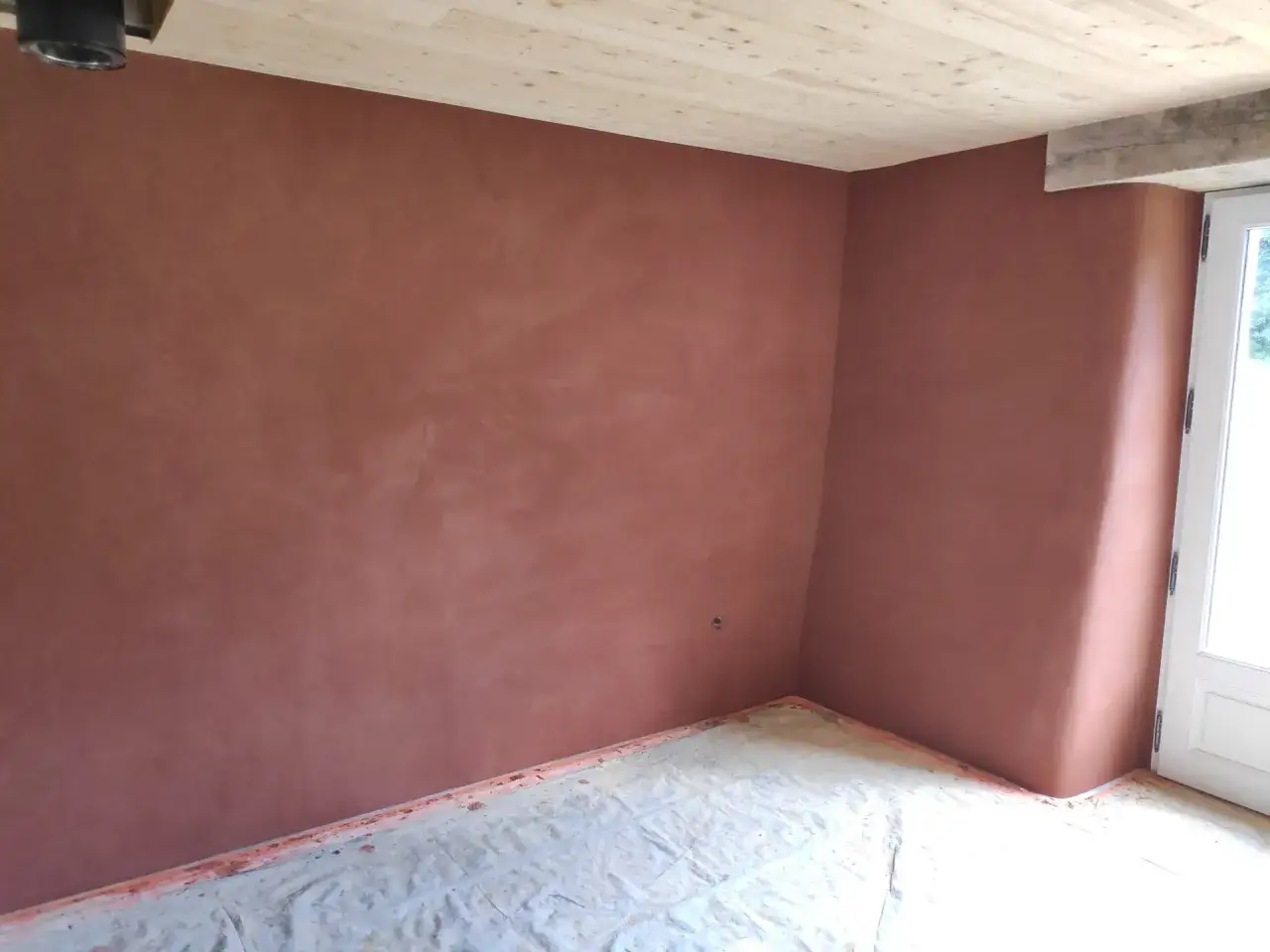 Un coin de pièce aux murs recouverts d'un enduit chaux finition terre cuite, le sol protégé par une bâche.