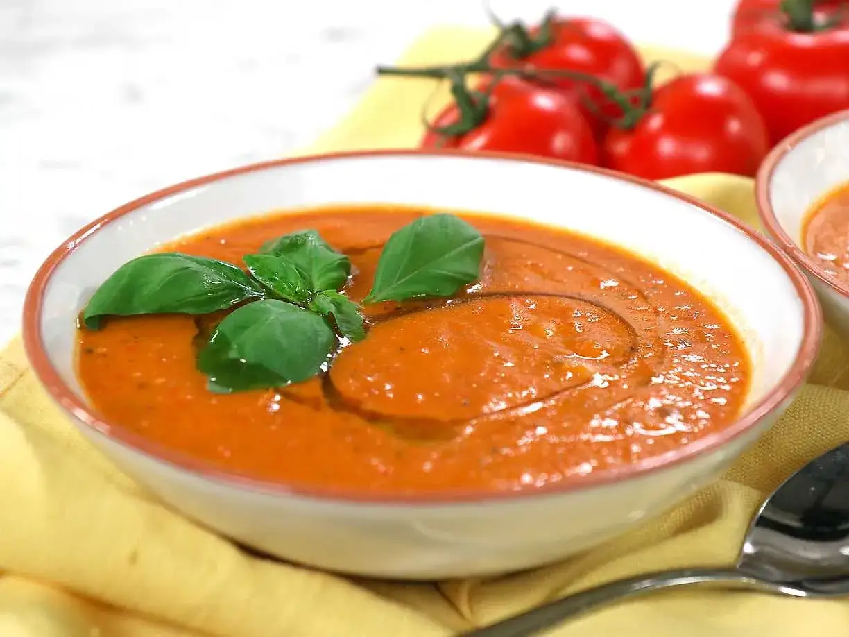 En skål med krämig rostad tomatsoppa, garnerad med färsk basilika och en virvel av olivolja. Tomater i bakgrunden.