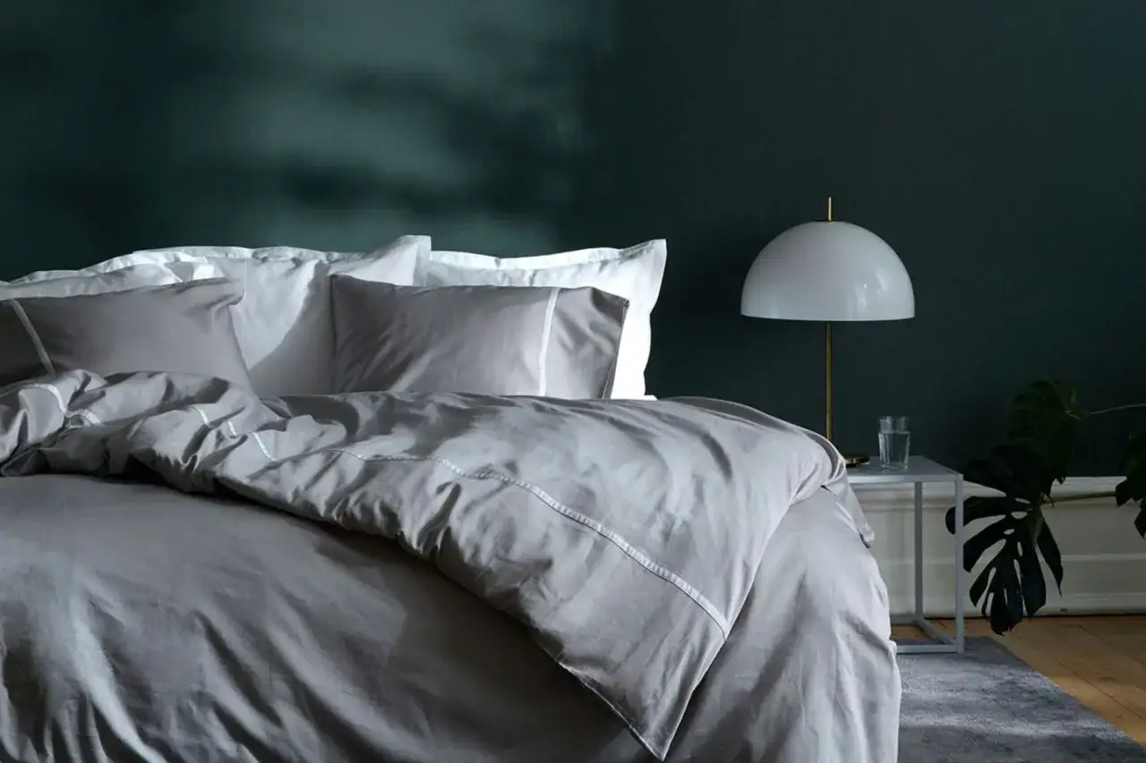 Ein gem&uuml;tliches Schlafzimmer mit dunkler Wandfarbe. Das Bett ist mit grauer Bettw&auml;sche und wei&szlig;en Kissen bezogen. Eine Lampe steht auf einem Nachttisch.