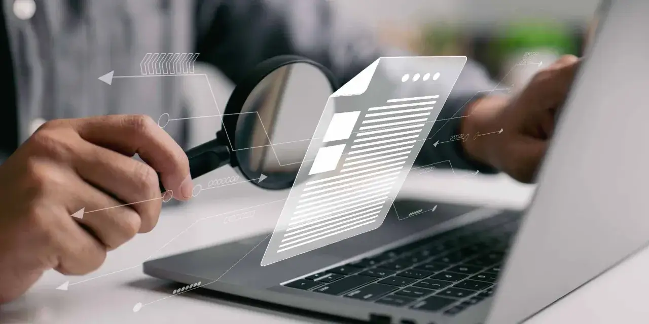 Ręka z lupą analizuje dokument na laptopie. Jak sprawdzić duplicate content?
