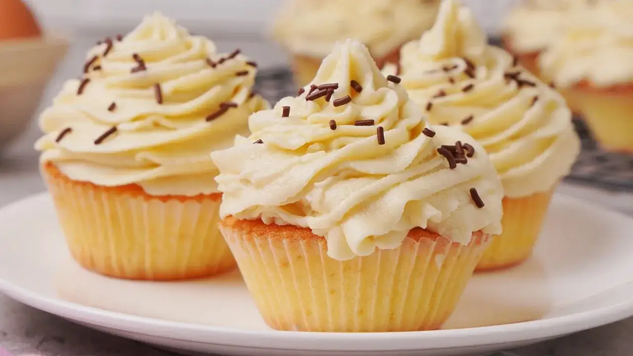 Mehrere k&ouml;stliche Vanille-Cupcakes mit cremiger Glasur und Schokostreuseln auf einem Teller.