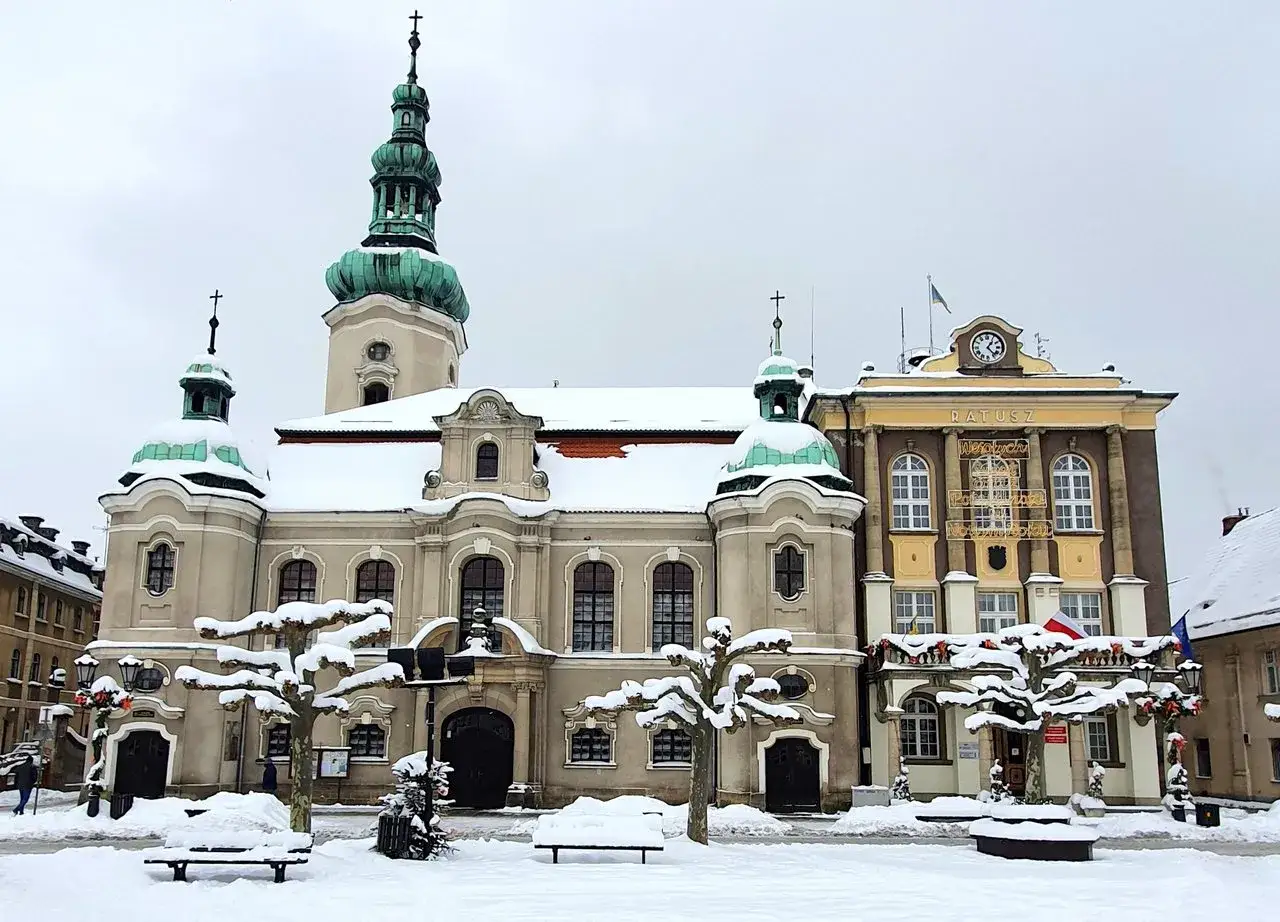 Zimowy rynek w Ćwiklicach. Śnieg pokrywa ławki, drzewa i dachy zabytkowych budynków, w tym ratusza.