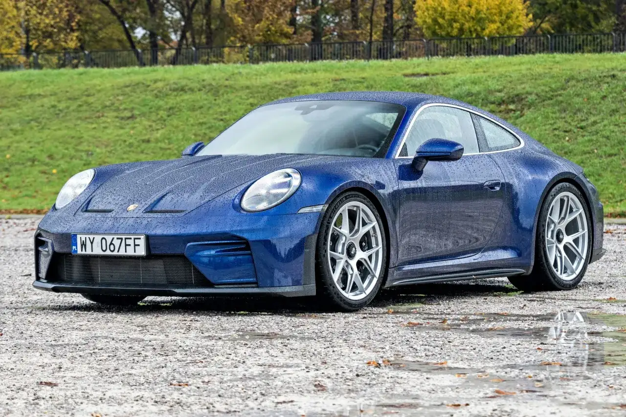 Niebieski Porsche 911 Carrera w deszczu, z kroplami wody na karoserii, stoi na mokrym żwirze.