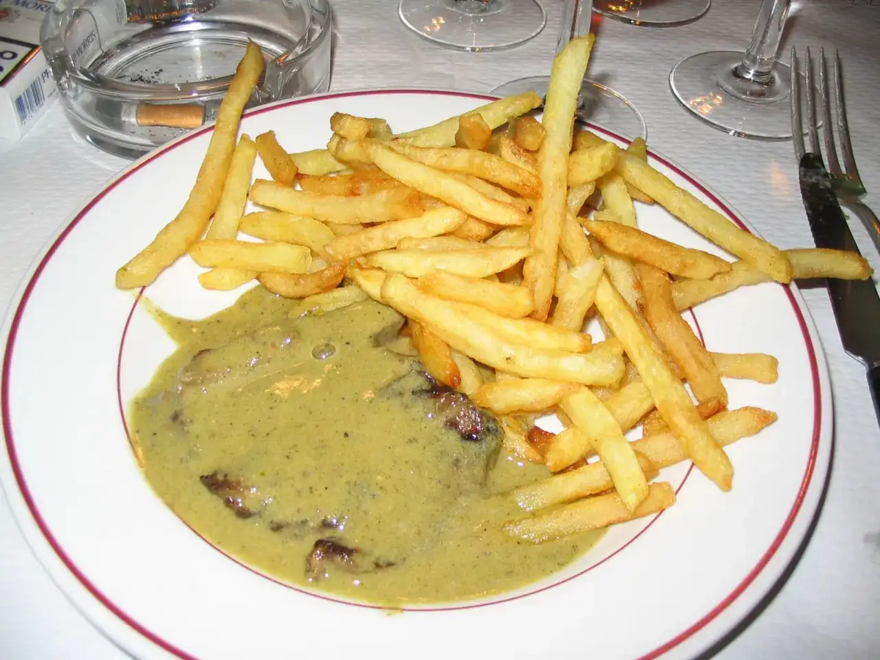 Ein Teller mit Steak und Pommes Frites, übergossen mit einer cremigen cafe de paris sauce. Daneben ein Aschenbecher mit Zigarettenstummel.