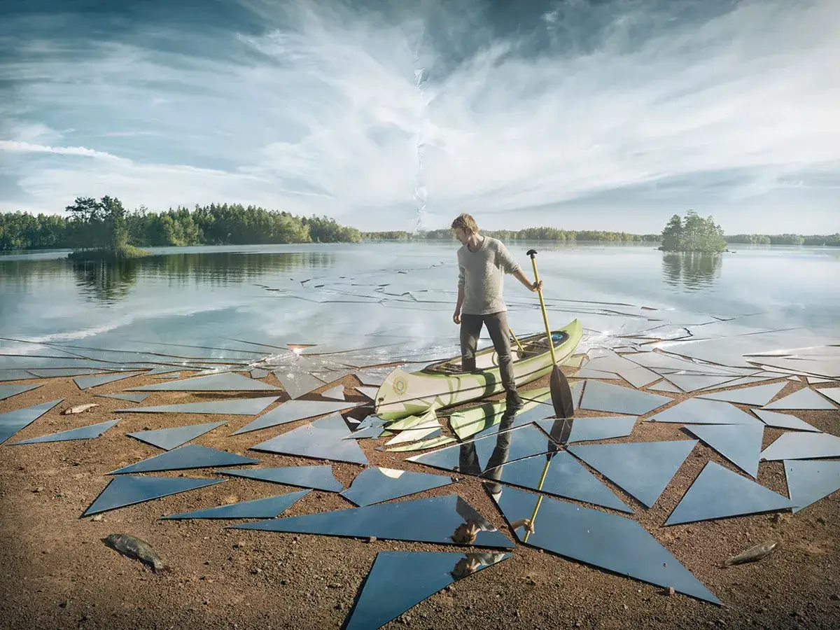 Un homme dans un canoë sur un lac gelé, entouré de morceaux de miroir brisés. La photo reflet crée une scène surréaliste.