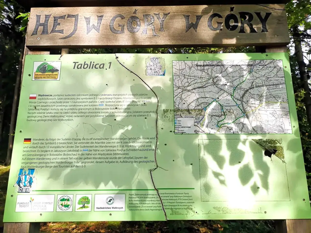 Tablica informacyjna o szlaku E-3 i geologii Gór Wałbrzyskich, z mapą i tekstem. W tle widoczny szczyt Chełmiec.