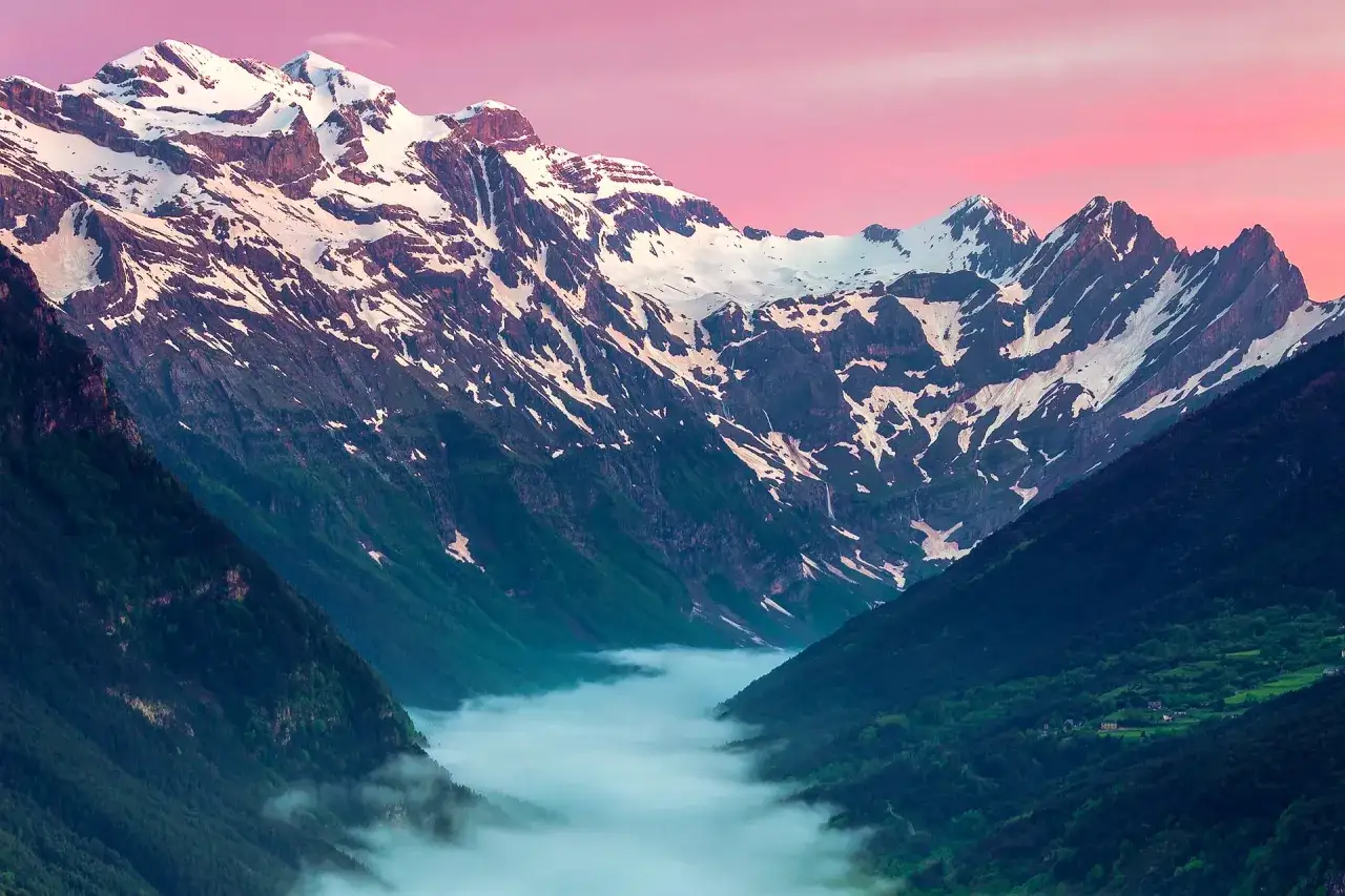 Majestuosas montañas nevadas se alzan sobre el **valle Pineta**, cubierto por una bruma matutina y exuberante vegetación verde.