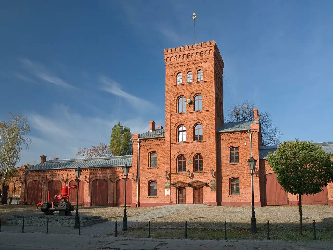 Stary budynek straży pożarnej, przypominający zamek, z wieżą i bramami garażowymi. Obok czerwona pompa, jakby z łodzi.