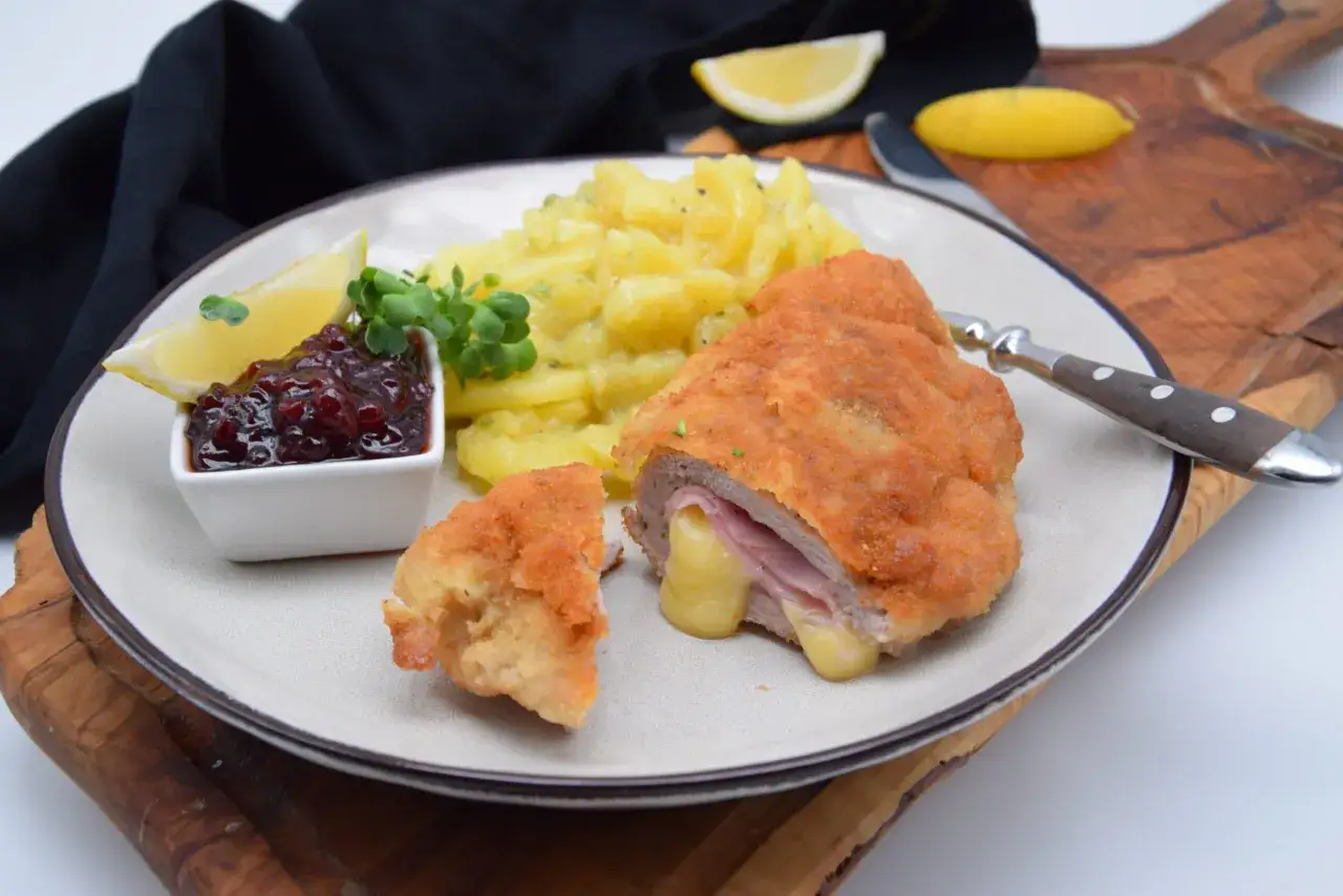 Ein knuspriges Cordon Bleu mit geschmolzenem K&auml;se, dazu Kartoffelsalat, Preiselbeeren und Zitrone auf einem Teller.