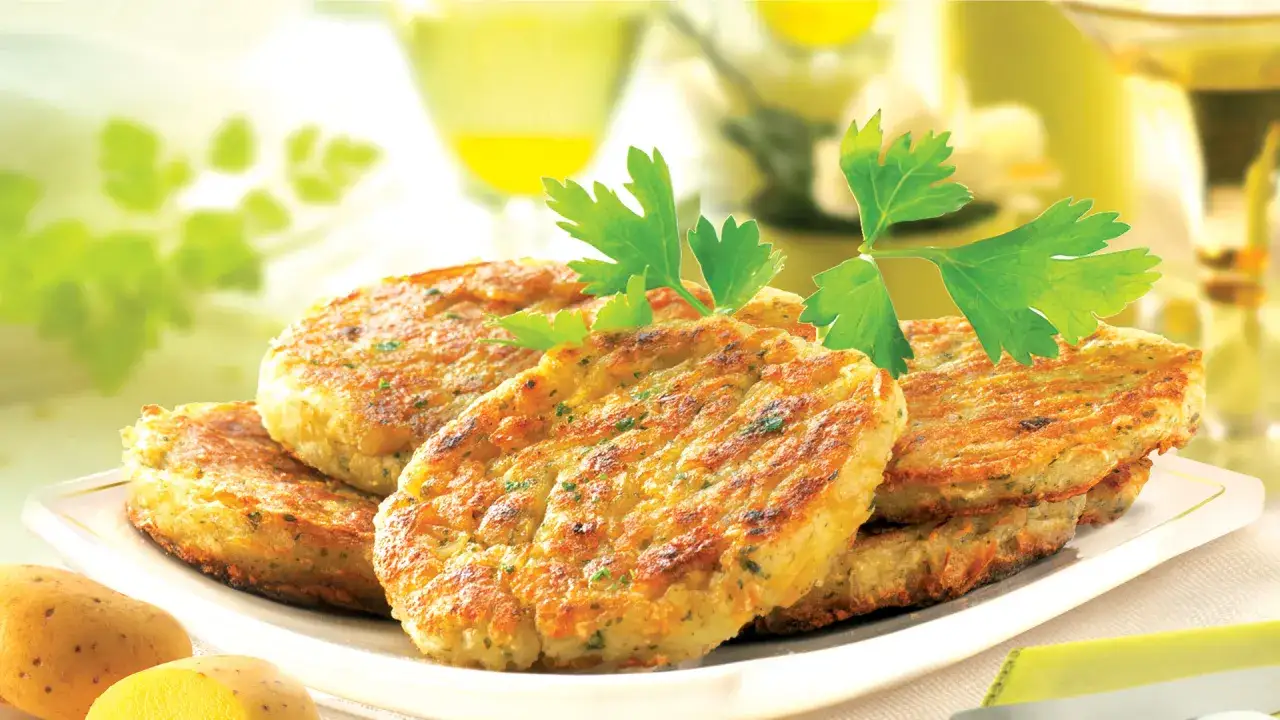 Pile de galettes de pomme de terre dor&eacute;es, garnies de persil frais, sur une assiette blanche. Des pommes de terre crues sont visibles au premier plan.