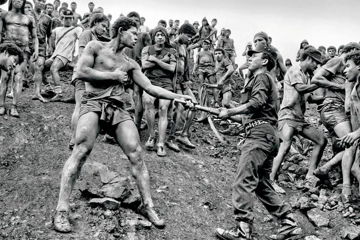 Sebastiao Salgado uchwycił moment, gdy dwaj mężczyźni w kopalni złota wymieniają się narzędziami. Wok&oacute;ł nich tłum innych g&oacute;rnik&oacute;w.