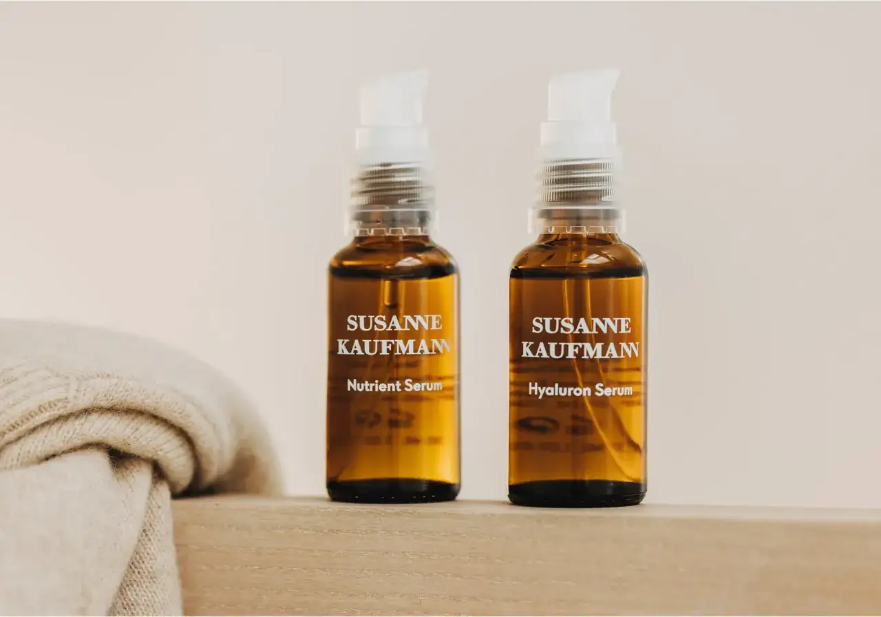 Zwei Fläschchen Susanne Kaufmann Hyaluron Serum und Nutrient Serum, ideal für die Gesichtspflege. Daneben liegt ein weicher Strickschal.
