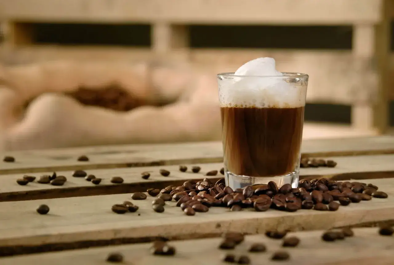 En caff&egrave; macchiato med fluffigt skum st&aring;r p&aring; tr&auml;bordet, omgiven av rostade kaffeb&ouml;nor.
