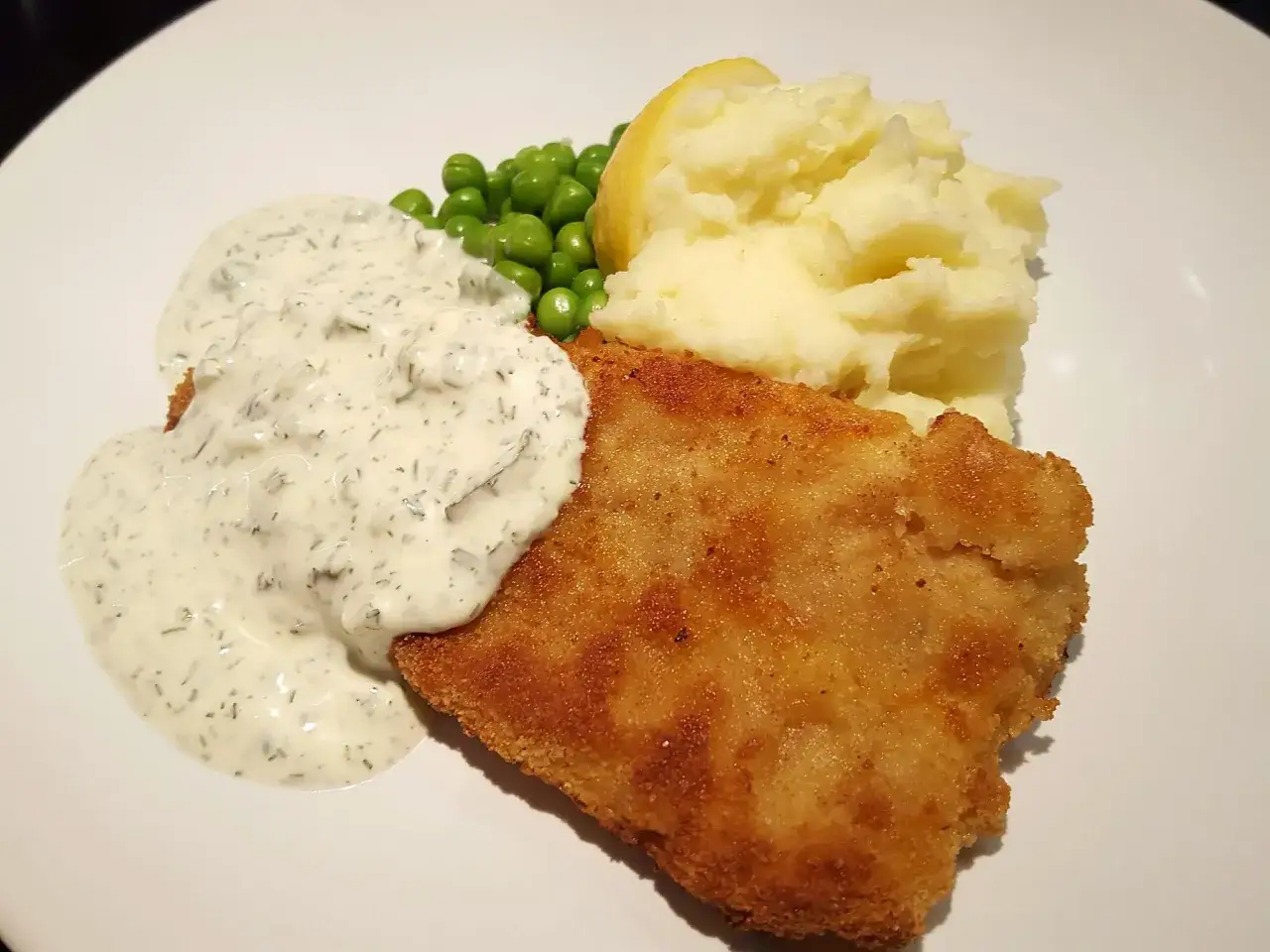 En gyllene panerad fiskfilé serveras med krämig remouladsås, fluffigt potatismos, gröna ärtor och en citronklyfta.
