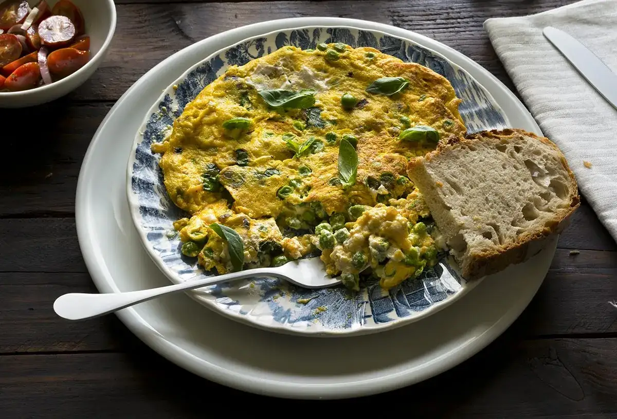 Tortilla de guisantes, esponjosa y dorada, servida con pan rústico y una ensalada de tomates.