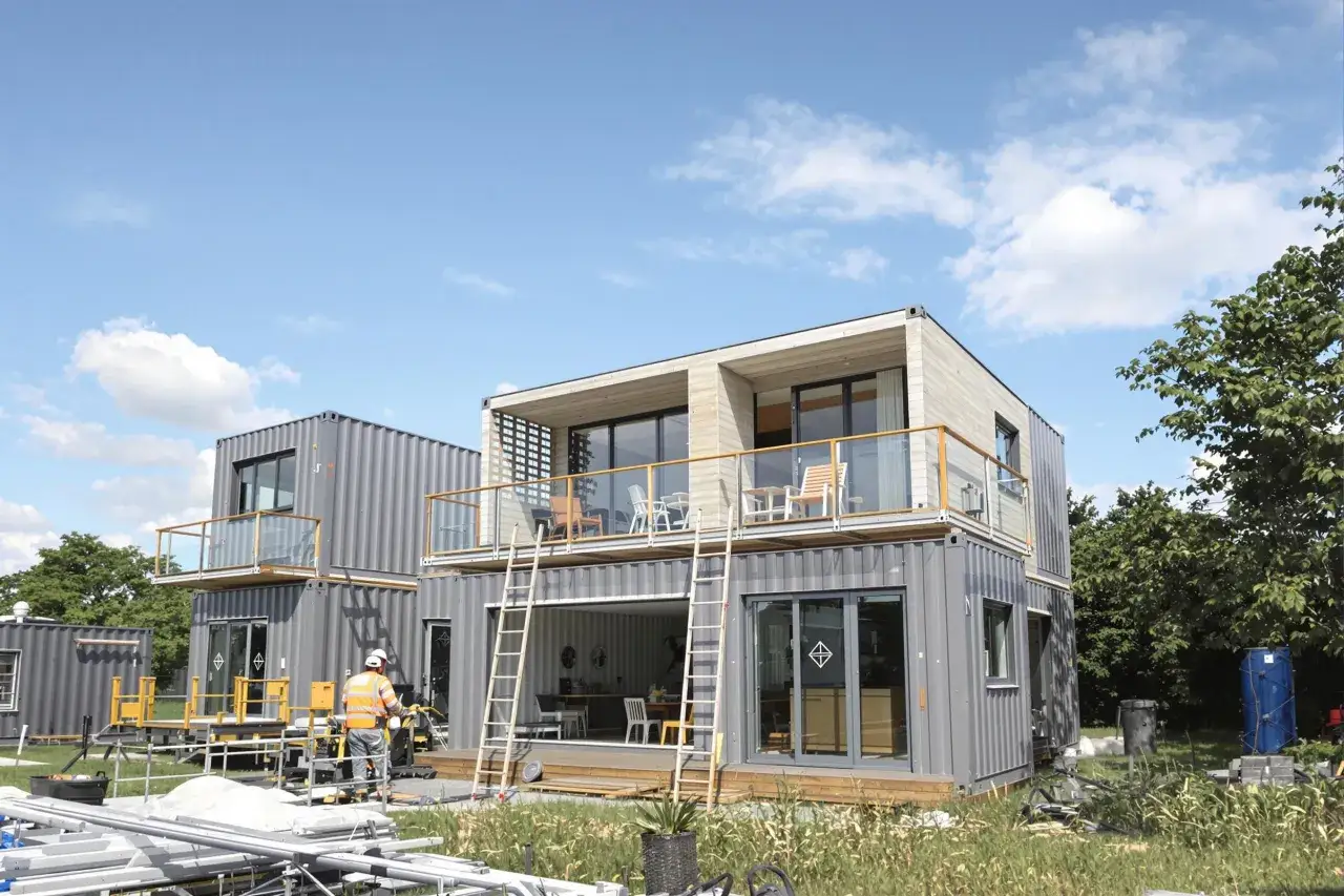 Maison en container prix : construction moderne &agrave; deux &eacute;tages avec balcons, fen&ecirc;tres panoramiques et un ouvrier en gilet orange.
