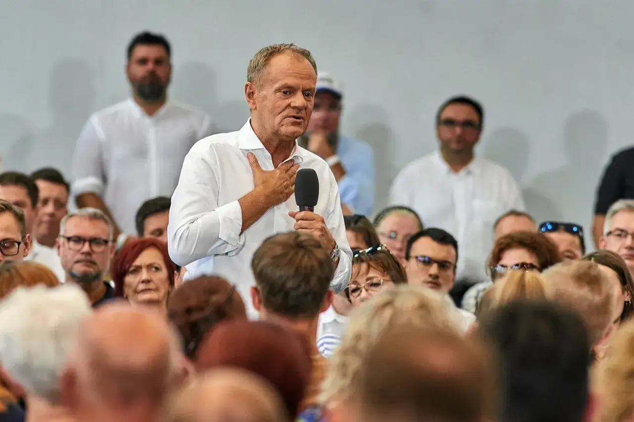 Donald Tusk przemawia do tłumu w Płocku, trzymając mikrofon i kładąc dłoń na sercu.