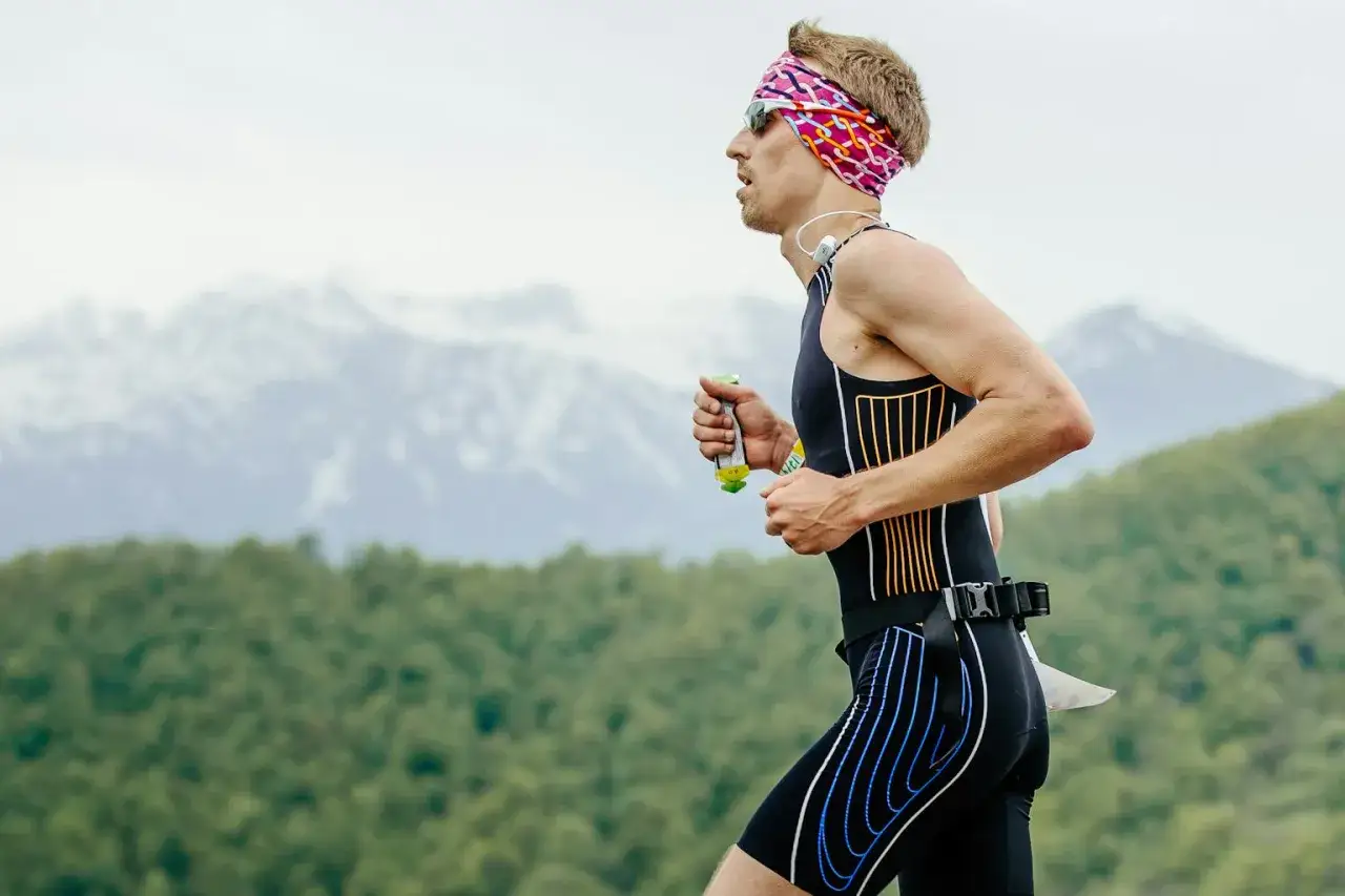 Zawodnik triathlonowy biegnie, trzymając w dłoni żel energetyczny. W tle góry i las.