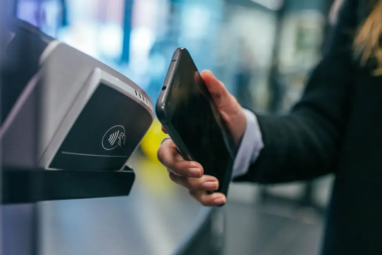 Dłoń zbliża telefon do terminala płatniczego z symbolem NFC. Szybkie płatności zbliżeniowe, czyli NFC co to jest, stają się codziennością.