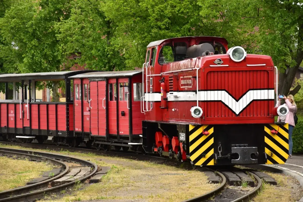 Czerwona lokomotywa z wagonami na torach, jedna z atrakcji Pałuk. Kobieta robi zdjęcie pociągowi.