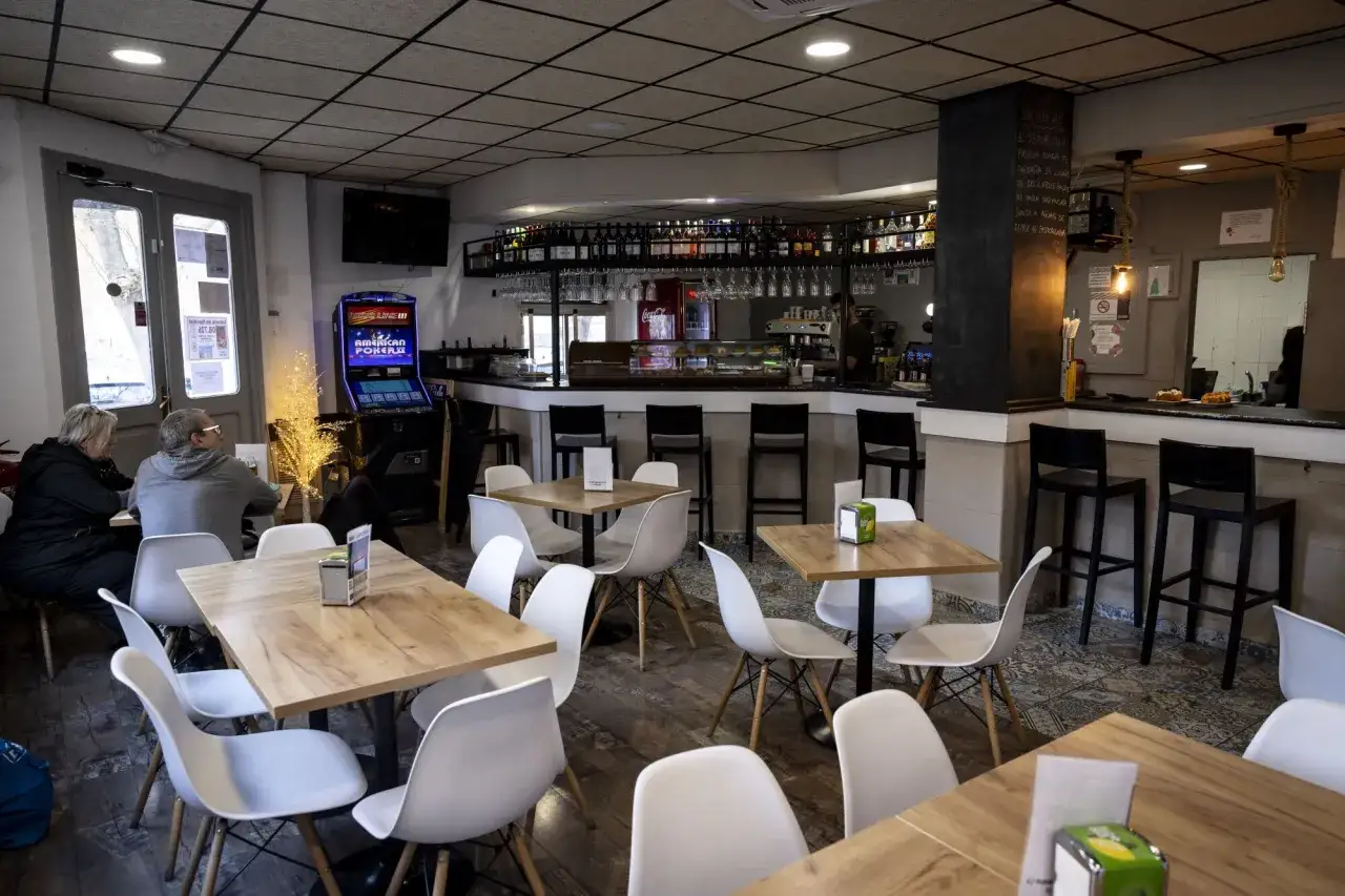 Interior de un bar con mesas y sillas, ideal para saber donde comer en el Actur. Hay una máquina de póker y una barra con bebidas.