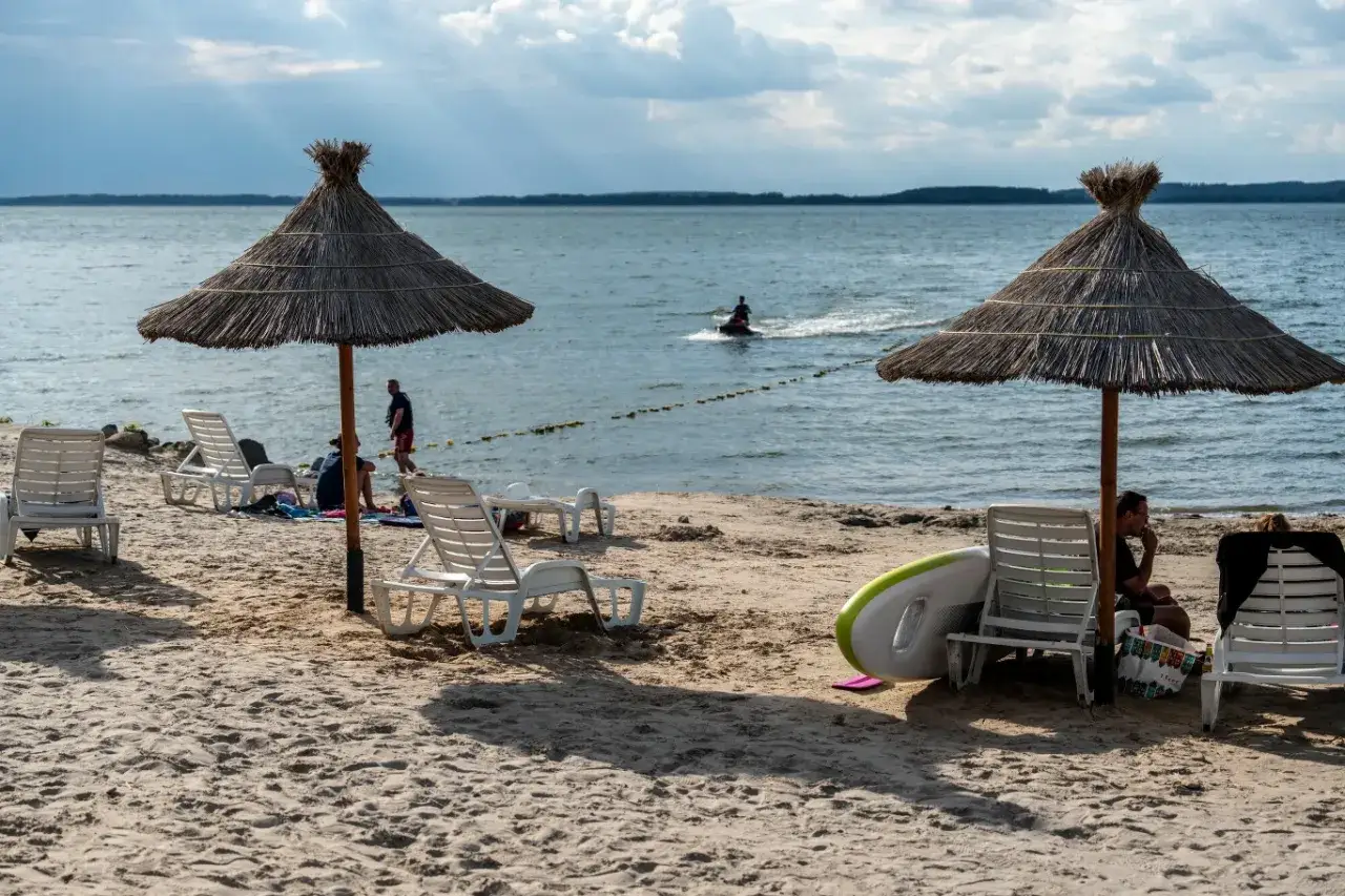 Słoneczny dzień na jednej z najpiękniejszych plaż w Polsce. Leżaki pod strzechą, woda i skuter wodny w oddali.