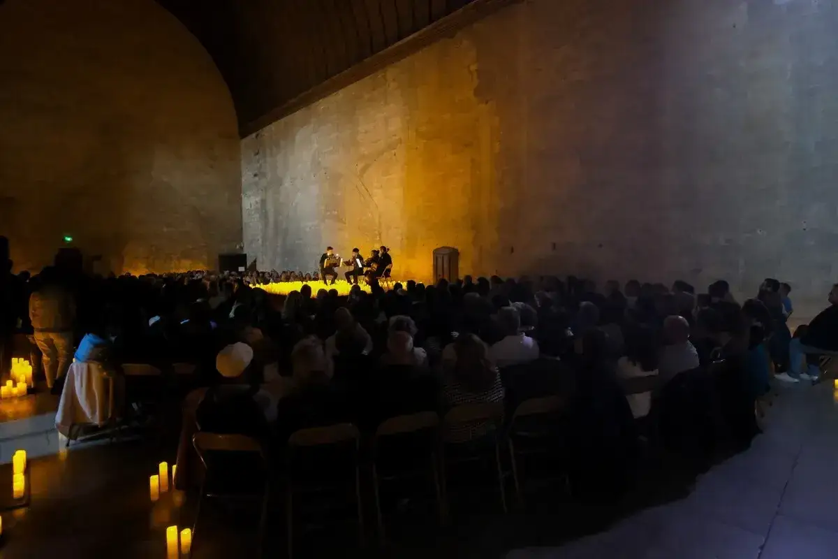 Concert aux chandelles dans un lieu historique pour une expo avignon. Musiciens sur sc&egrave;ne, public assis dans la p&eacute;nombre.