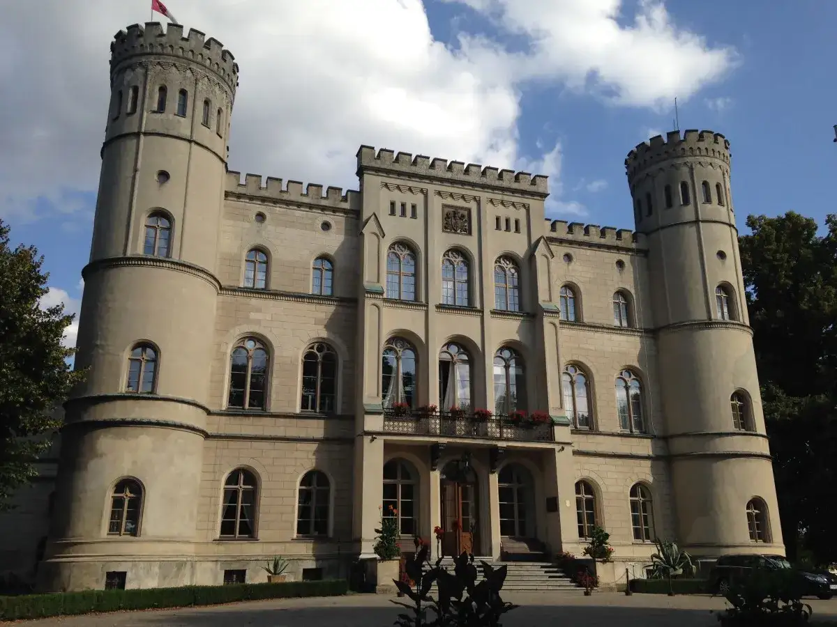 Zamek w Rokosowie, z dwiema okrągłymi wieżami i blankami, prezentuje się majestatycznie na tle błękitnego nieba z chmurami.
