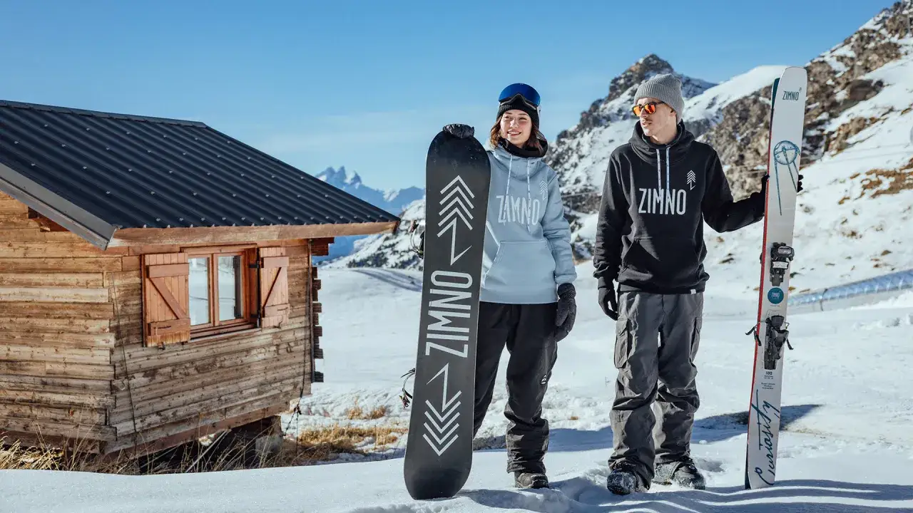 Para na tle górskiej chatki prezentuje stylowy ubiór na narty i snowboard. Ona w jasnej bluzie, on w ciemnej.