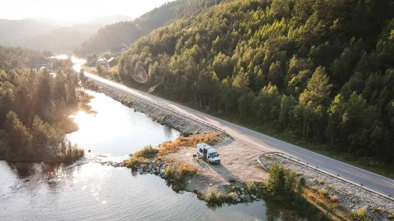 Ein Wohnmobil parkt am Ufer eines Flusses an einer Stra&szlig;e in Schweden. Die Sonne scheint durch die B&auml;ume und wirft lange Schatten.