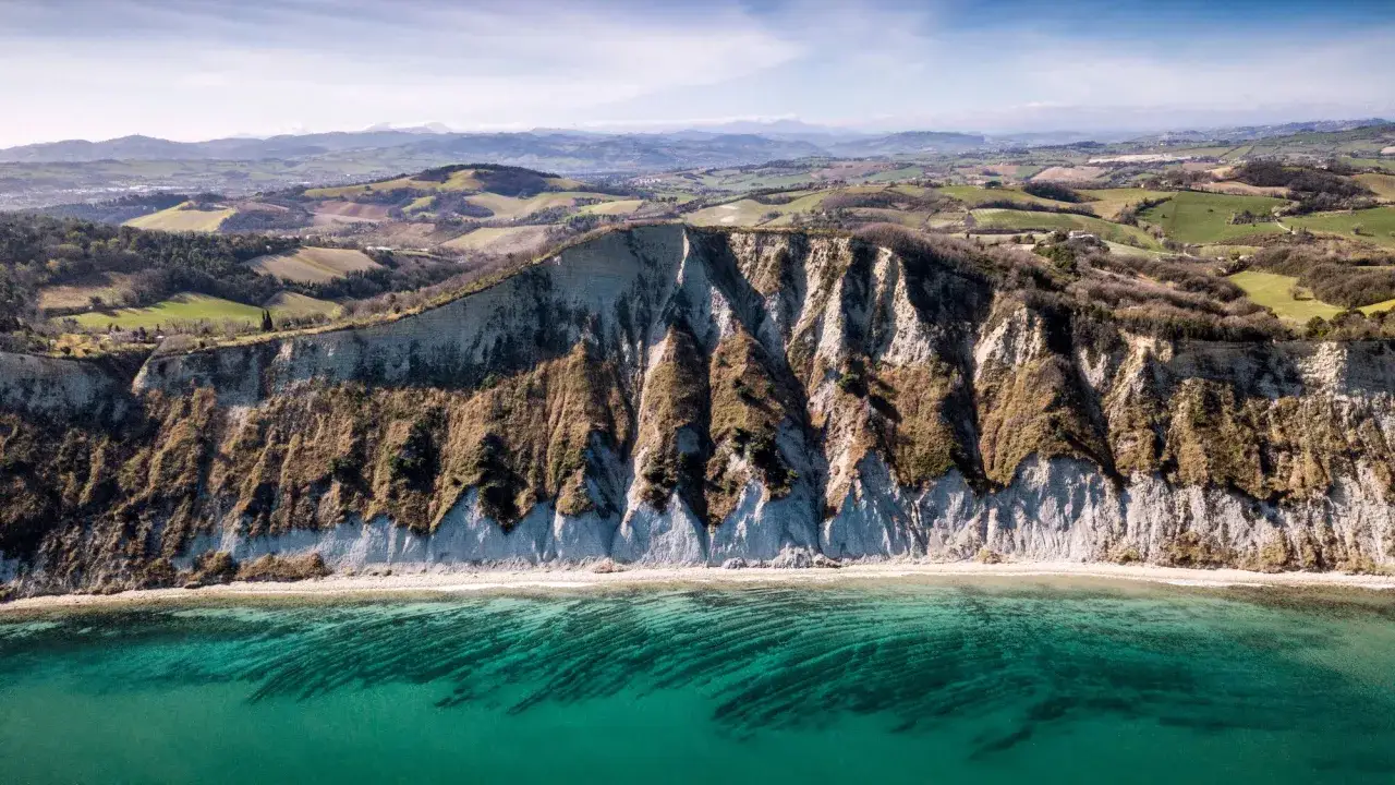 Scogliere a picco sul mare turchese nel parco naturale regionale del Monte San Bartolo. Colline verdi e campi coltivati sullo sfondo.