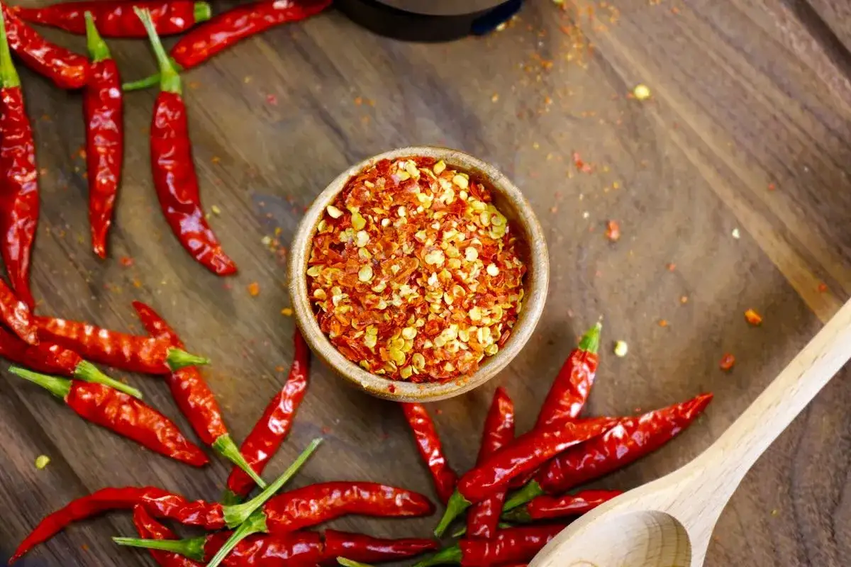 Rote Chilischoten, aus denen Cayennepfeffer hergestellt wird, liegen auf einem Holzbrett. Eine Schale mit zersto&szlig;enen Chilis steht daneben.