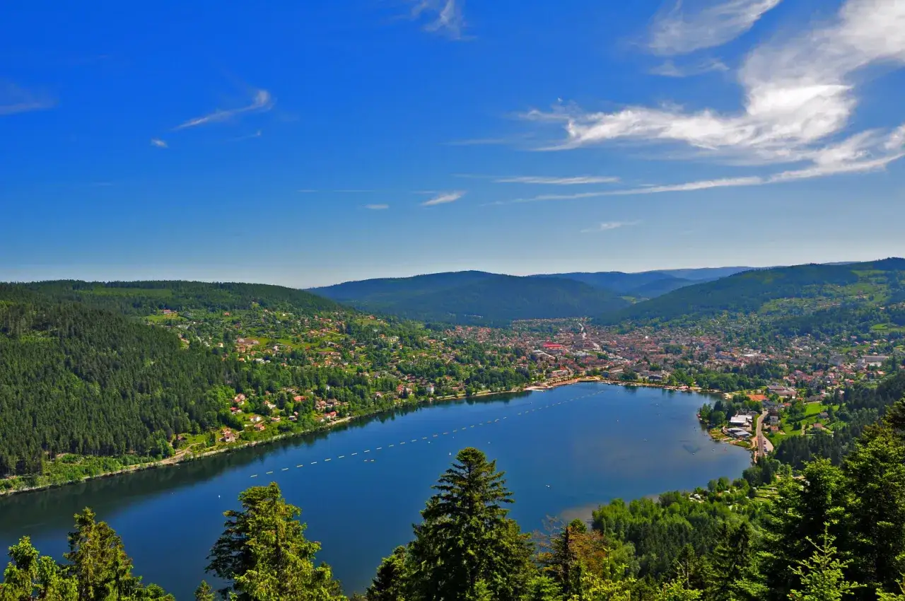Vue panoramique du lac de Gérardmer, entouré de forêts verdoyantes et de maisons pittoresques, sous un ciel bleu éclatant.