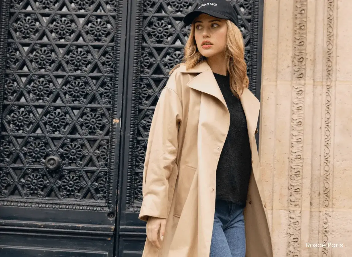 Une **look femme 40 ans moderne** d&eacute;contract&eacute;e, portant un trench beige, un pull noir et un jean, devant une porte ornement&eacute;e.