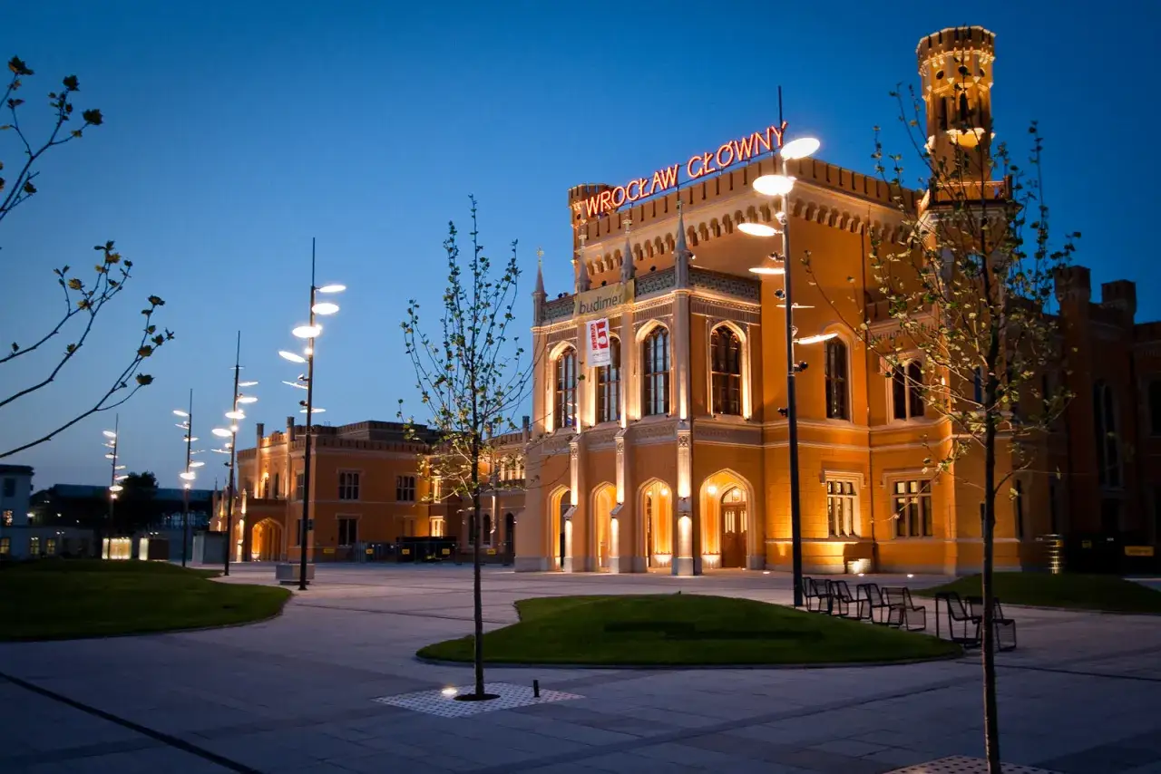 Der beleuchtete Bahnhof Breslau erstrahlt im Abendlicht. Moderne Laternen s&auml;umen den Platz vor dem historischen Geb&auml;ude.