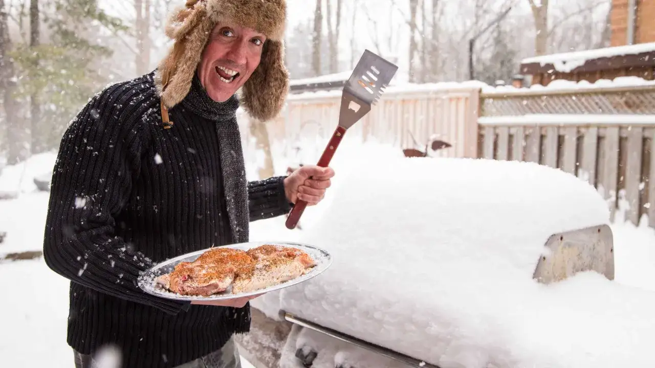 Un homme en bonnet de fourrure prépare un barbecue d'hiver, tenant une assiette de viande assaisonnée et une spatule.