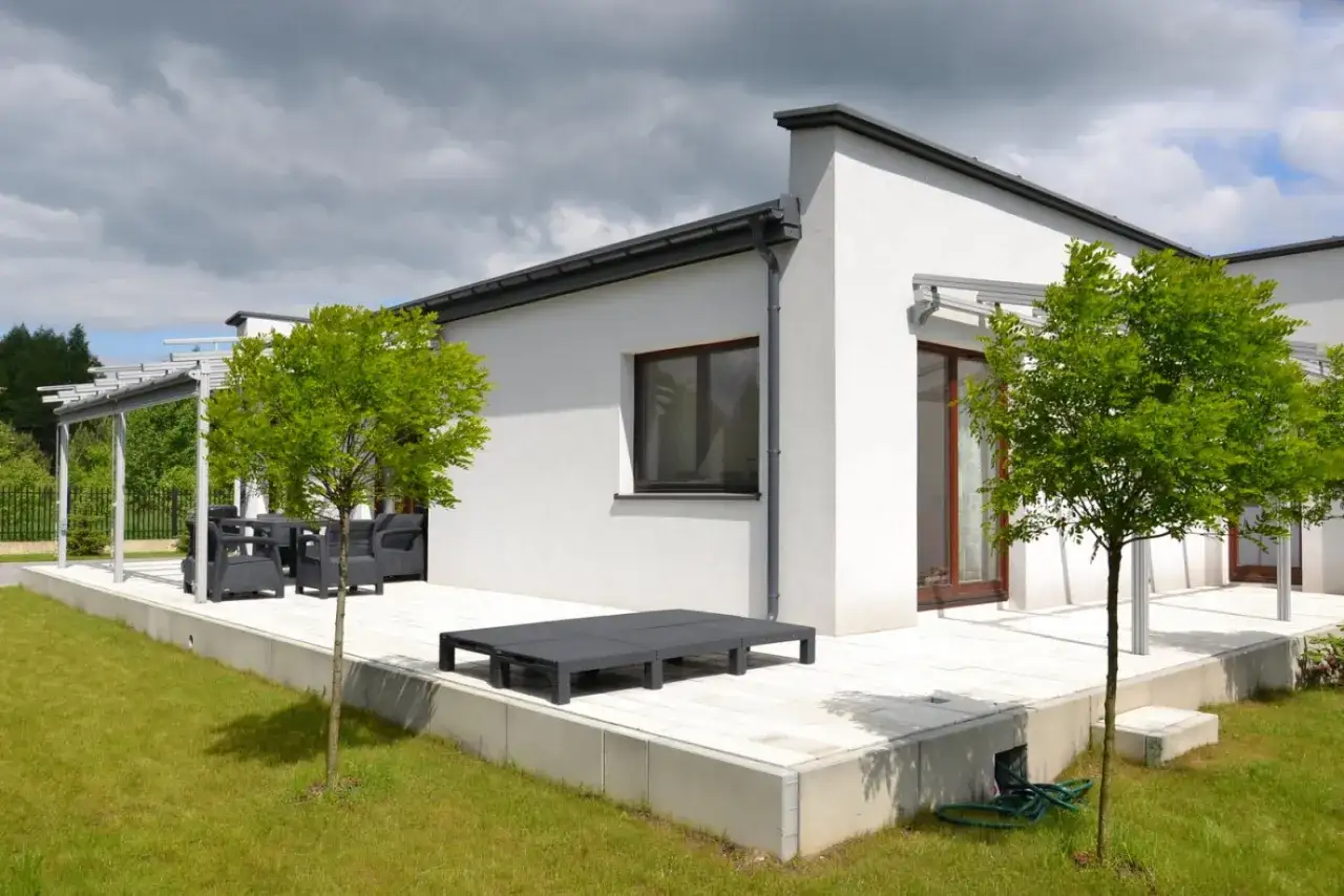 Nowoczesny bungalow z tarasem i meblami ogrodowymi. Obok rosną młode drzewka.