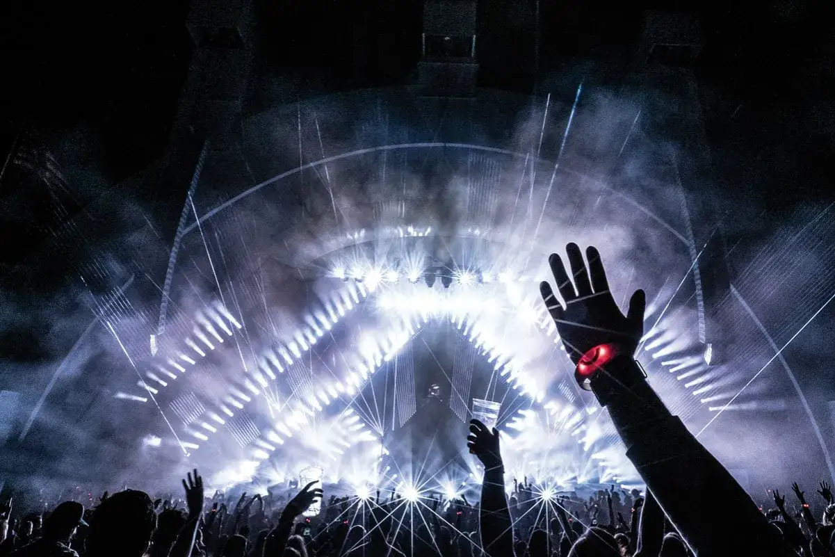 Eine Hand hebt sich im Takt der EDM-Musik. Die Bühne erstrahlt in hellem Licht und Nebel, die Menge feiert.
